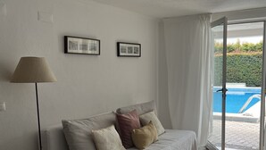 A modern living room with a sofa, a lamp, a small table with a plant, and a view of a swimming pool through a glass door.
