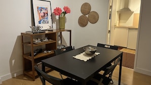 A dining area with a black table, chairs, a framed picture, a vase with flowers, and a shelf with dishes and a kettle.