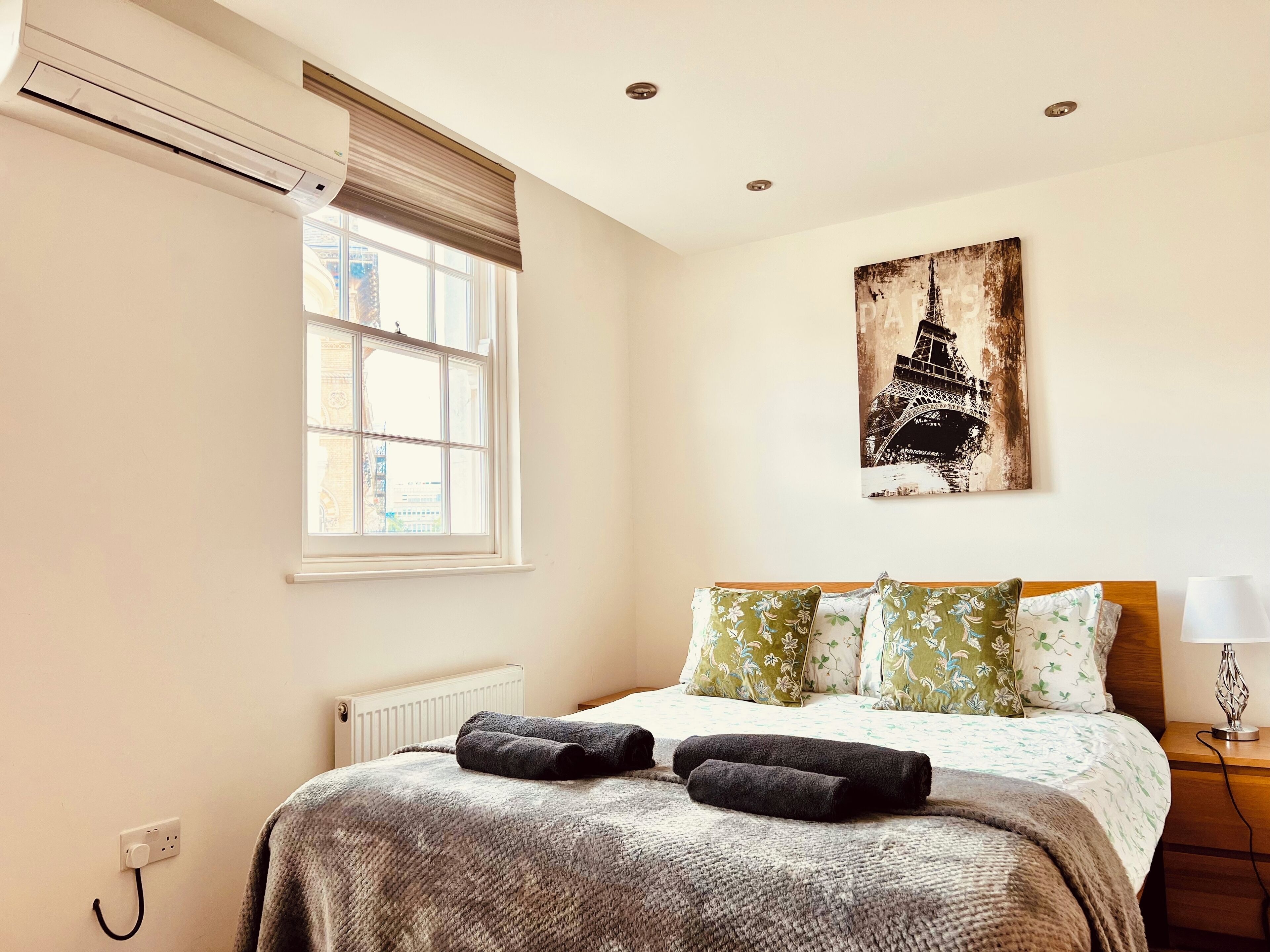 A bedroom with a bed, a radiator, a window with blinds, a wall-mounted air conditioner, a lamp, and a painting of the Eiffel Tower.