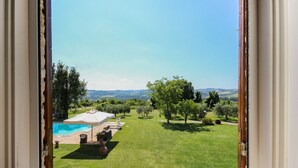 Vista dalla finestra su un giardino con piscina, patio con mobili da esterno e prato verde con alberi.