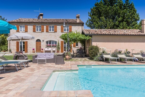 A pool area with lounge chairs, a table with chairs, and a building with a tiled roof and stone walls. - Villa Panorama by Anita Villas (Serrungarina)