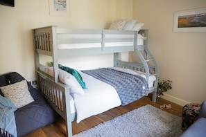 Ein Etagenbett mit weißer Matratze, einer grauen Decke und gemusterten Kissen. Auf dem Boden liegt ein grauer Teppich und neben dem Bett steht eine kleine Pflanze.