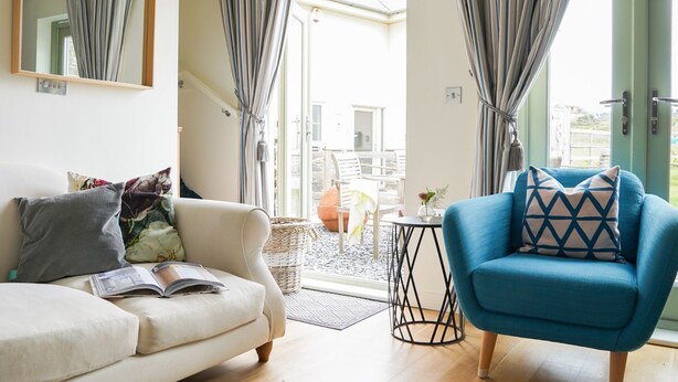 Ein Wohnzimmer mit einer Couch, einem blauen Sessel und einem kleinen Tisch. Vor den Fenstern hängen Vorhänge und an der Wand befindet sich ein Spiegel.