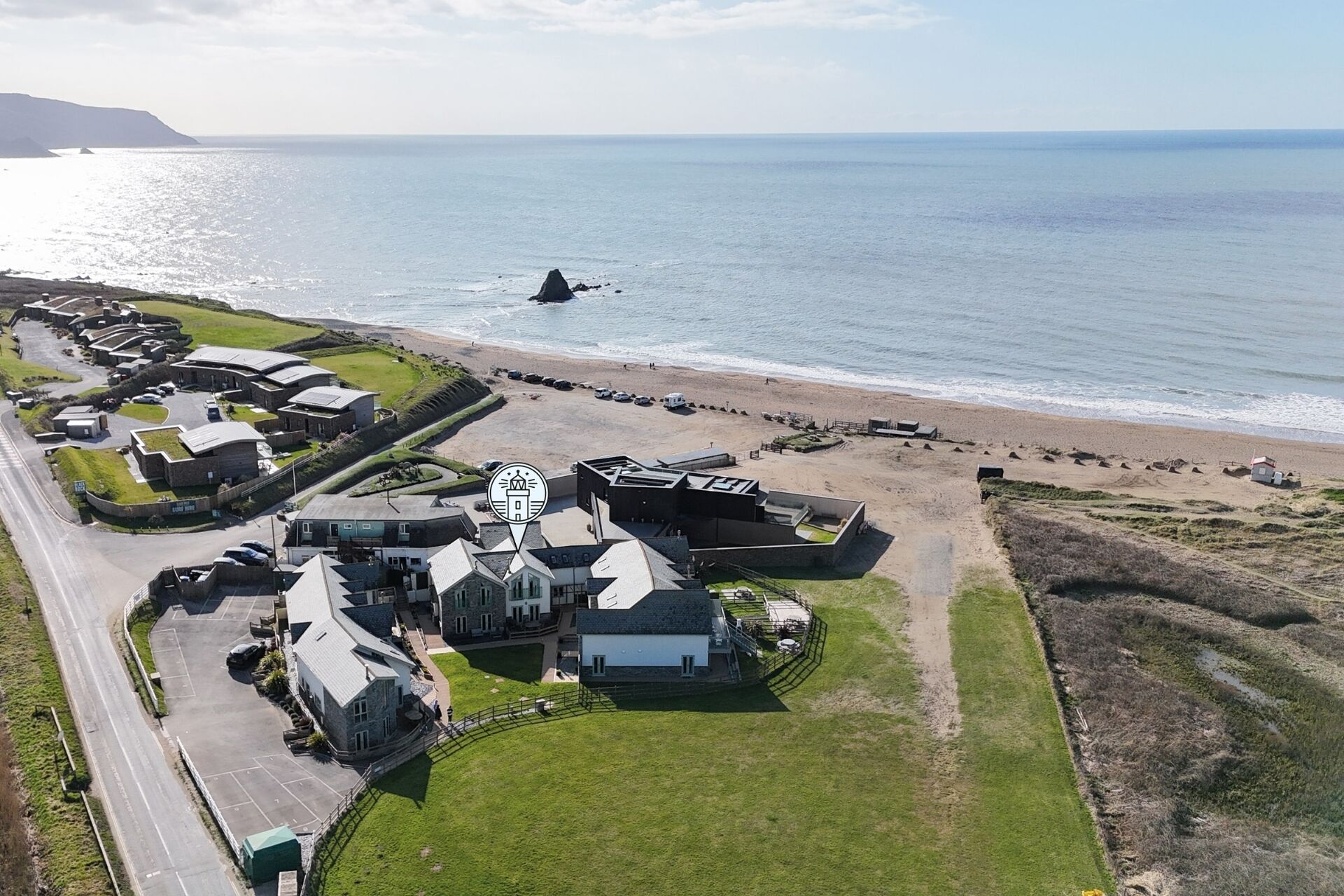 Vue aérienne d’une propriété située sur la côte, comprenant plusieurs bâtiments, un espace de stationnement et une plage.