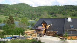 Una cabaña de madera con techo inclinado, un pequeño porche y vista a un lago y colinas boscosas.