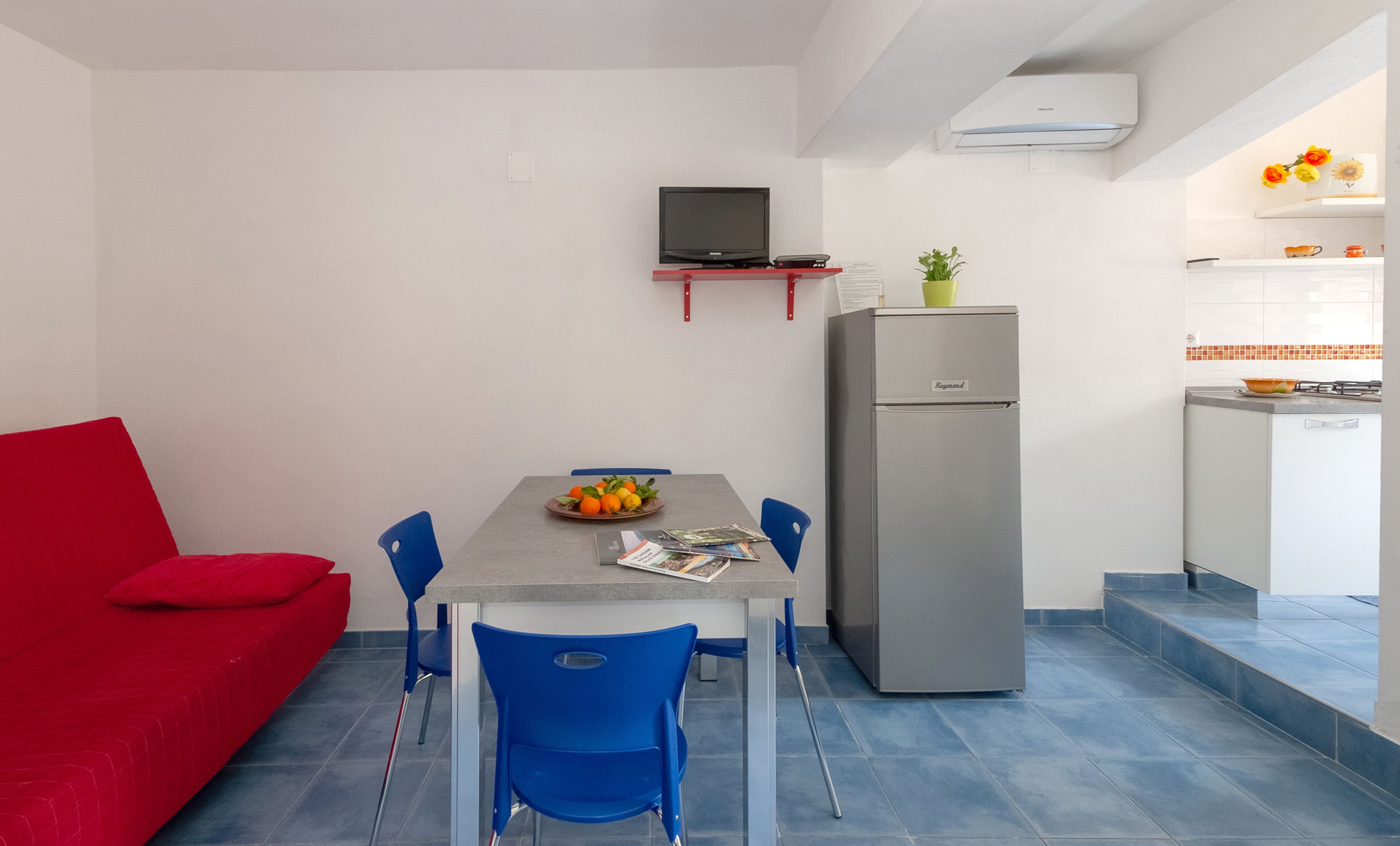 Un espace de vie compact comprenant un coin repas, un canapé rouge, des chaises bleues, un réfrigérateur gris, une petite cuisine avec une cuisinière et un téléviseur à écran plat fixé au mur.
