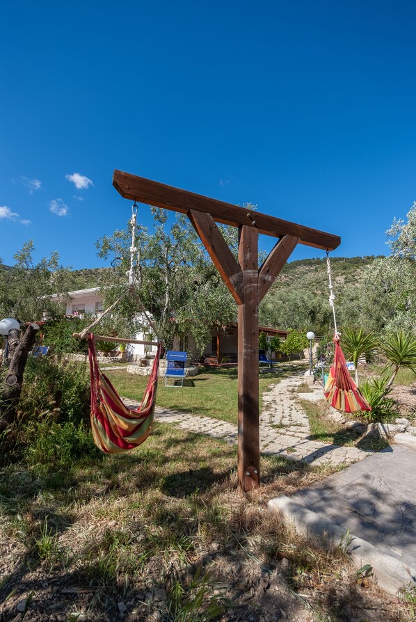 A wooden pergola with two hammocks hanging from it, set in a garden with a clear blue sky and a path leading to a building in the background.
