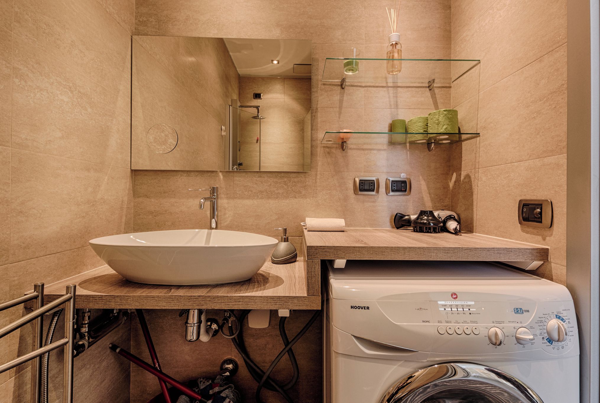 Ein modernes Badezimmer mit einem großen Waschbecken, einem Spiegel, einer Glasablage für Pflegeprodukte und einer Waschmaschine.