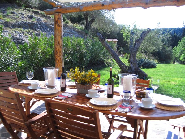 An outdoor dining area with wooden tables and chairs, surrounded by greenery and a view of the countryside.
