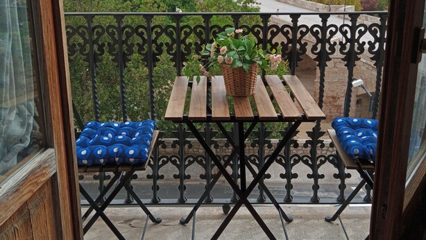 Un balcón con una mesa, dos sillas, una planta en maceta y vistas de una ciudad con edificios y zonas verdes.