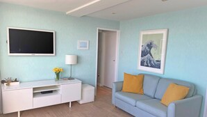 A modern living room with a flat-screen TV, a white entertainment unit, a grey sofa with yellow cushions, and a framed artwork on the wall. - Stunning Seafront Penthouse w/ Private Sun Terrace & Direct Panoramic Sea Views (Kings Road)