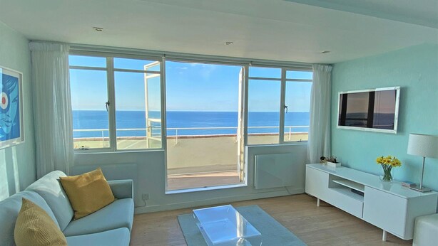 A modern living room with a large window overlooking the beach, a sofa, a coffee table, a TV, and a small plant. - Stunning Seafront Penthouse w/ Private Sun Terrace & Direct Panoramic Sea Views (Kings Road)