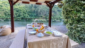 A table set outdoors with a view of a lake and forest, covered with a beige tablecloth, and various food items and drinks.