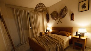 Un dormitorio con cama de madera, dos mesitas de noche con lámparas, un ventanal con cortinas y adornos en la pared, incluyendo un abanico y una máscara.