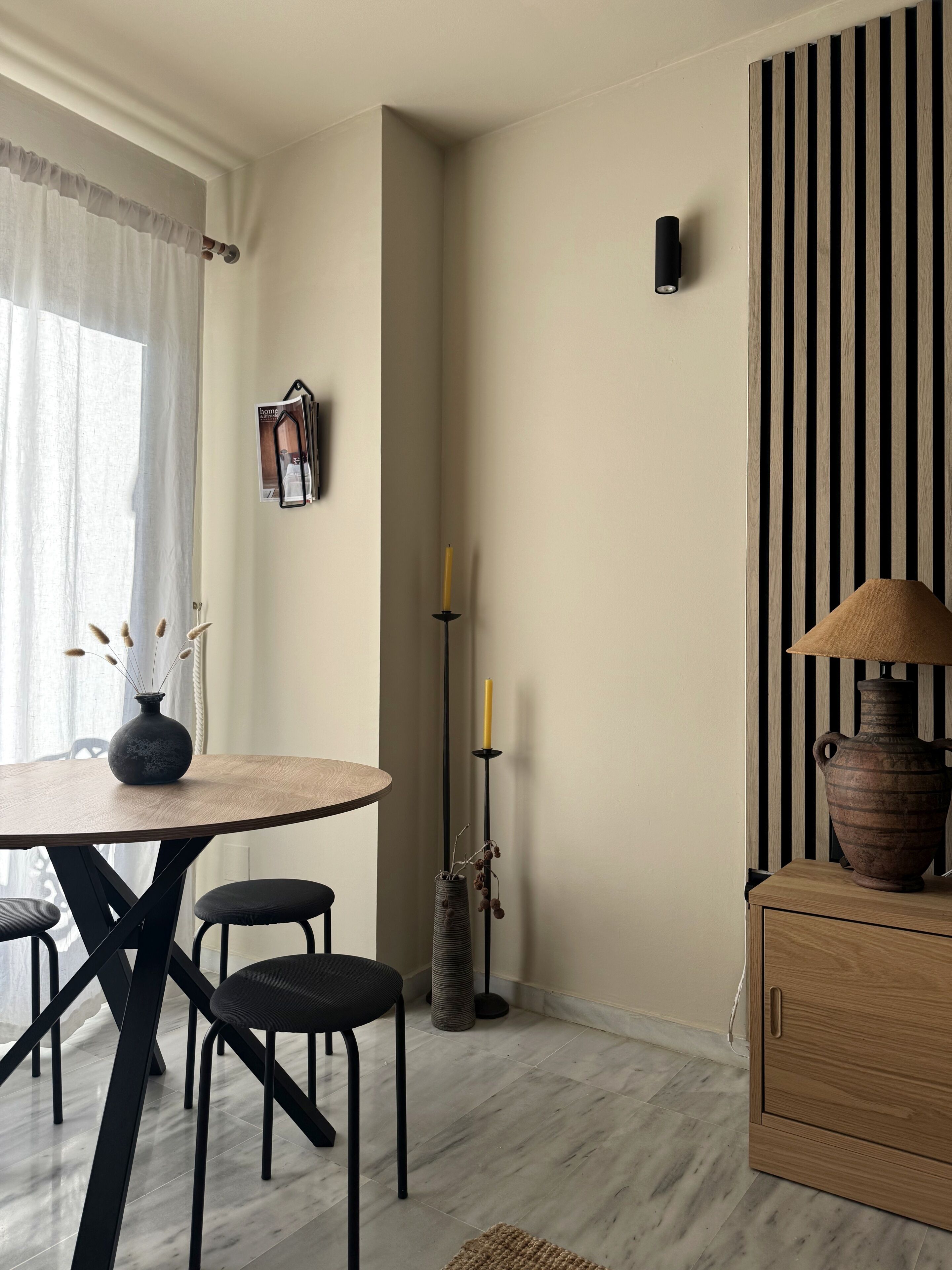 Een kamer met een ronde tafel, twee stoelen, een vaas met takken, een hoge kandelaar, een lamp op een kast en een aan de muur hangend schilderij.