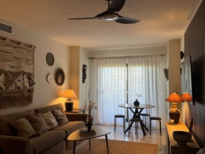 A living room with a sofa, coffee table, and a dining table with chairs. There is a ceiling fan, a wall-mounted clock, and a window with curtains.