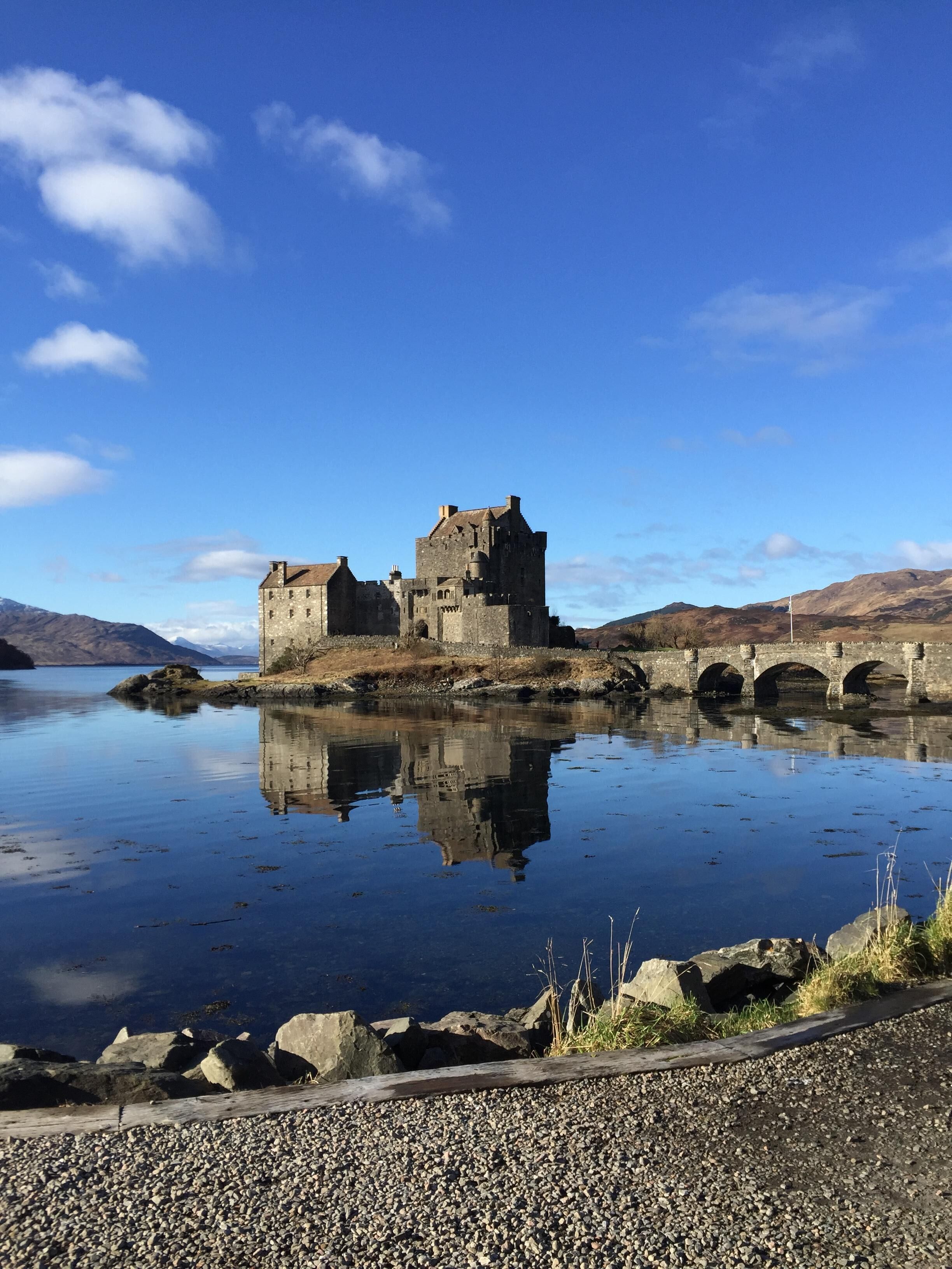 Un château de pierre chargé d’histoire, situé au bord d’un lac paisible, sous un ciel dégagé.
