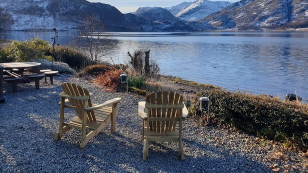 A lakeside seating area with two Adirondack chairs and a table, overlooking a calm lake with mountains in the background. - A loch side property with stunning views located in a walker's paradise. (Inverinate)