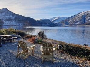Un área de estar junto al lago con dos sillas Adirondack y una mesa, con vistas a un lago tranquilo y montañas al fondo.