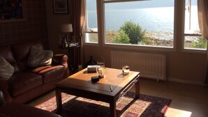 A living room with a brown leather sofa, a wooden coffee table, and a view of the sea through large windows. - A loch side property with stunning views located in a walker's paradise. (Inverinate)