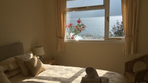 A neatly made bed with a rolled towel, a bedside lamp, and a view of the sea through a window with curtains. - A loch side property with stunning views located in a walker's paradise. (Inverinate)
