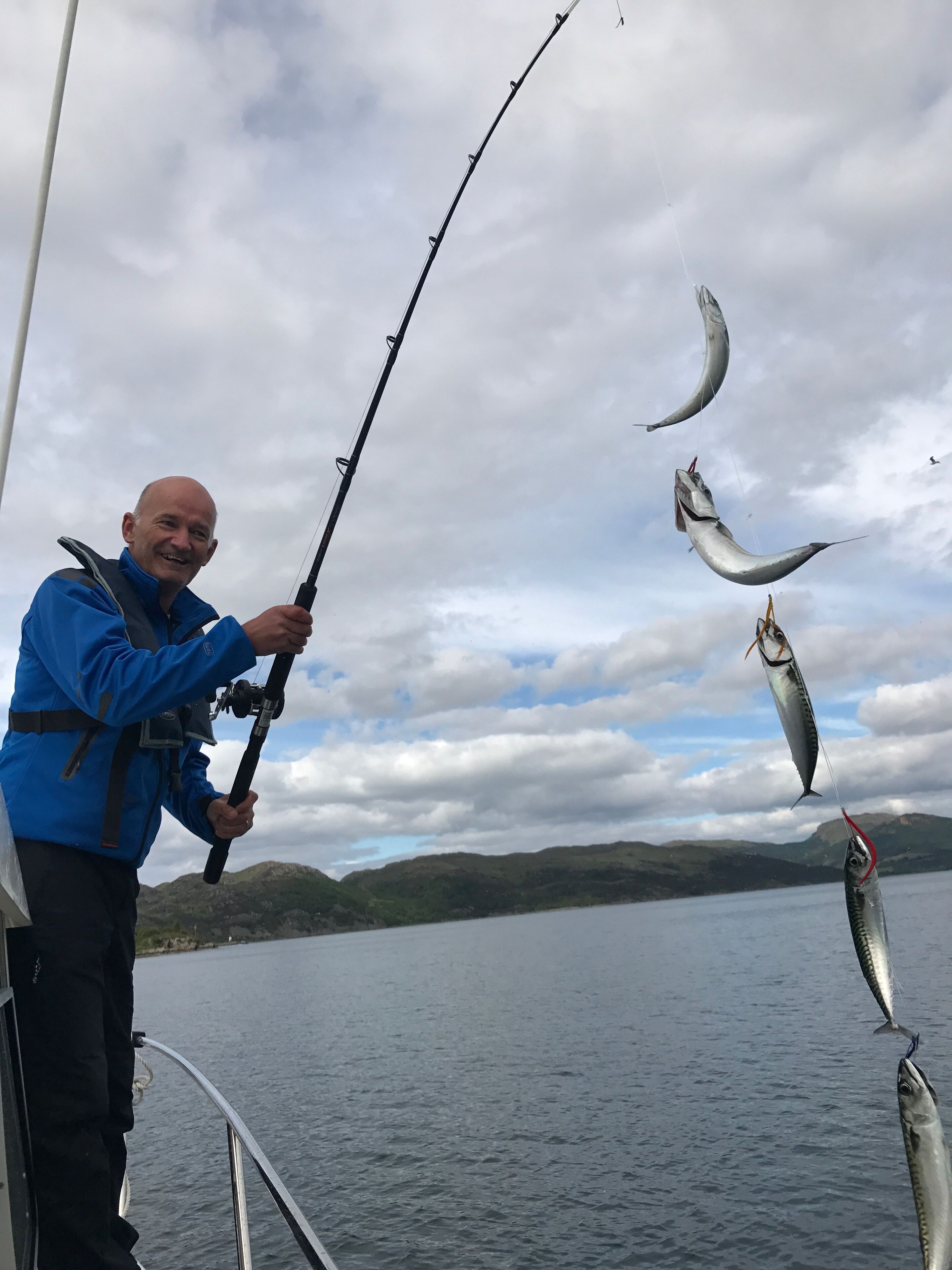 Una persona che tiene una canna da pesca con diversi pesci, in piedi su una barca, con un lago e delle colline sullo sfondo.