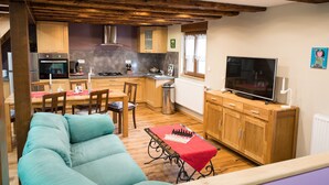 Una sala de estar acogedora con un sofá color turquesa, una mesa de comedor de madera con sillas, un televisor sobre un mueble de madera, y una zona de cocina con armarios de madera y una estufa.