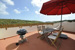 Een terras met een barbecue, een tafel en stoelen, met uitzicht op een waterpartij en heuvels.