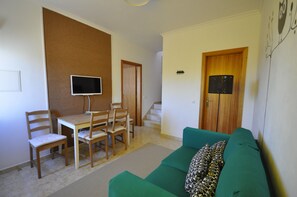 Een woonkamer met een groene bank, een houten eettafel en stoelen. Er is een televisie aan de muur bevestigd, een trap en een deur met een bordje.