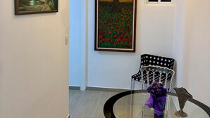 Un pasillo con un cuadro enmarco de un paisaje, una lámpara de araña, una silla y una mesa con un jarrón.