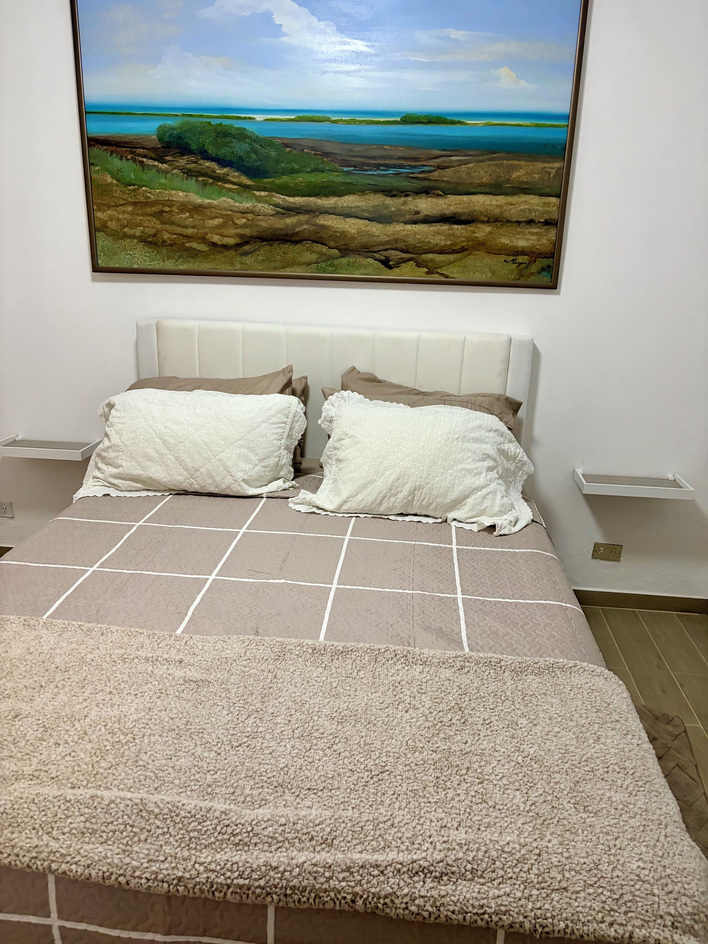 A bed with a quilt, pillows, and a view of a landscape painting.