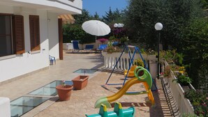 Un patio con zona de juegos infantiles, que incluye un columpio y un tobogán. Hay plantas en macetas y mobiliario exterior.
