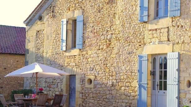 Un edificio de piedra con encanto rústico, que presenta un comedor con mesa y sillas, y una puerta con contraventanas azules.