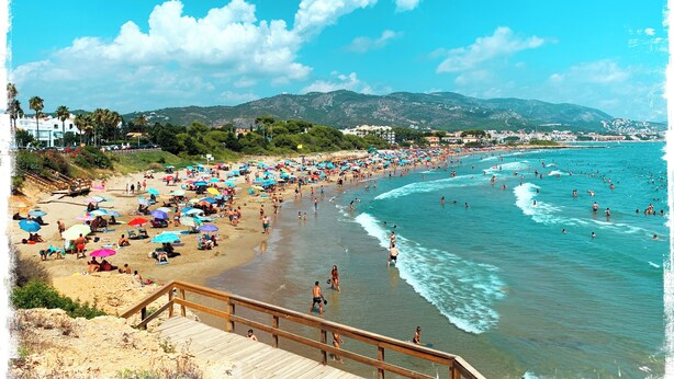 Una playa con un paseo de madera que lleva al agua, sombrillas y gente disfrutando del mar.