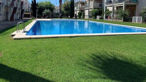 Una piscina rectangular rodeada de césped cuidado, con un edificio de varios pisos que tiene balcones y muebles de exterior al fondo.