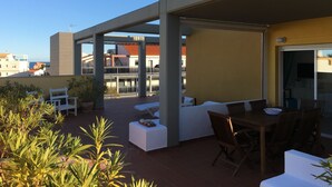 Un espacio al aire libre cubierto con bancos blancos acolchados y una mesa de madera, rodeado de plantas en macetas.