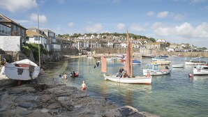 A coastal town with boats docked along the shore, a rocky beach, and buildings in the background. - Light filled home from home with spectacular panoramic sea views of Mounts Bay (Mousehole)