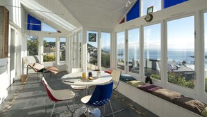A sunlit room with a dining table, chairs, and a bench along a large window overlooking the sea. - Light filled home from home with spectacular panoramic sea views of Mounts Bay (Mousehole)