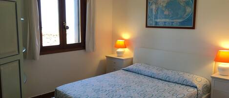 A bedroom with a bed, two bedside lamps, a window with curtains, a wall map, and a wardrobe.