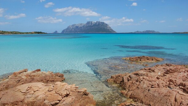 A coastal landscape with clear turquoise waters, rocky foreground, and a prominent mountain in the distance.