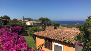 A small building with a tiled roof, surrounded by vibrant pink bougainvillea flowers, and a view of the sea and distant hills.