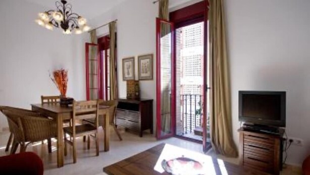 A room with a wooden dining table, chairs, a television on a wooden stand, and a balcony with a red door and curtains. - Beautiful apartment in  historical recently reformed building. (Sevilla)