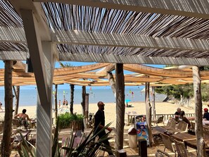 A beachside restaurant with outdoor seating, wooden pillars, and a view of the beach.