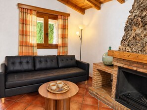 A living room with a black leather sofa, a round wooden coffee table with two cups and a tray, a fireplace with a stone mantle, and a window with plaid curtains.
