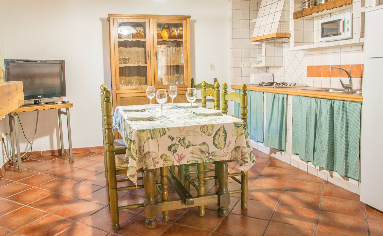 A dining area with a table set for four, a wooden cabinet with glass doors, a television on a stand, and a kitchen area with a microwave and sink.