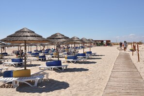 Ein Strand mit Liegestühlen, die unter Reetdächern in Reihen angeordnet sind, ein Holzsteg und ein klarer Himmel.