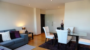 Een moderne woonkamer met een grijze bank, een glazen eettafel met witte stoelen en een donker vloerkleed.