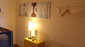 Een kamer met een bed, een nachtkastje met een lamp, een 'ENJOY' bord aan de muur en een kapstok met hangers.