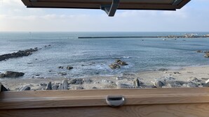 Vista desde una habitación con un amplio ventanal que da a una playa rocosa y al mar.