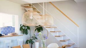 Una cocina moderna con comedor, que presenta una escalera de madera, lámparas colgantes de mimbre y un jarrón con flores sobre la mesa.
